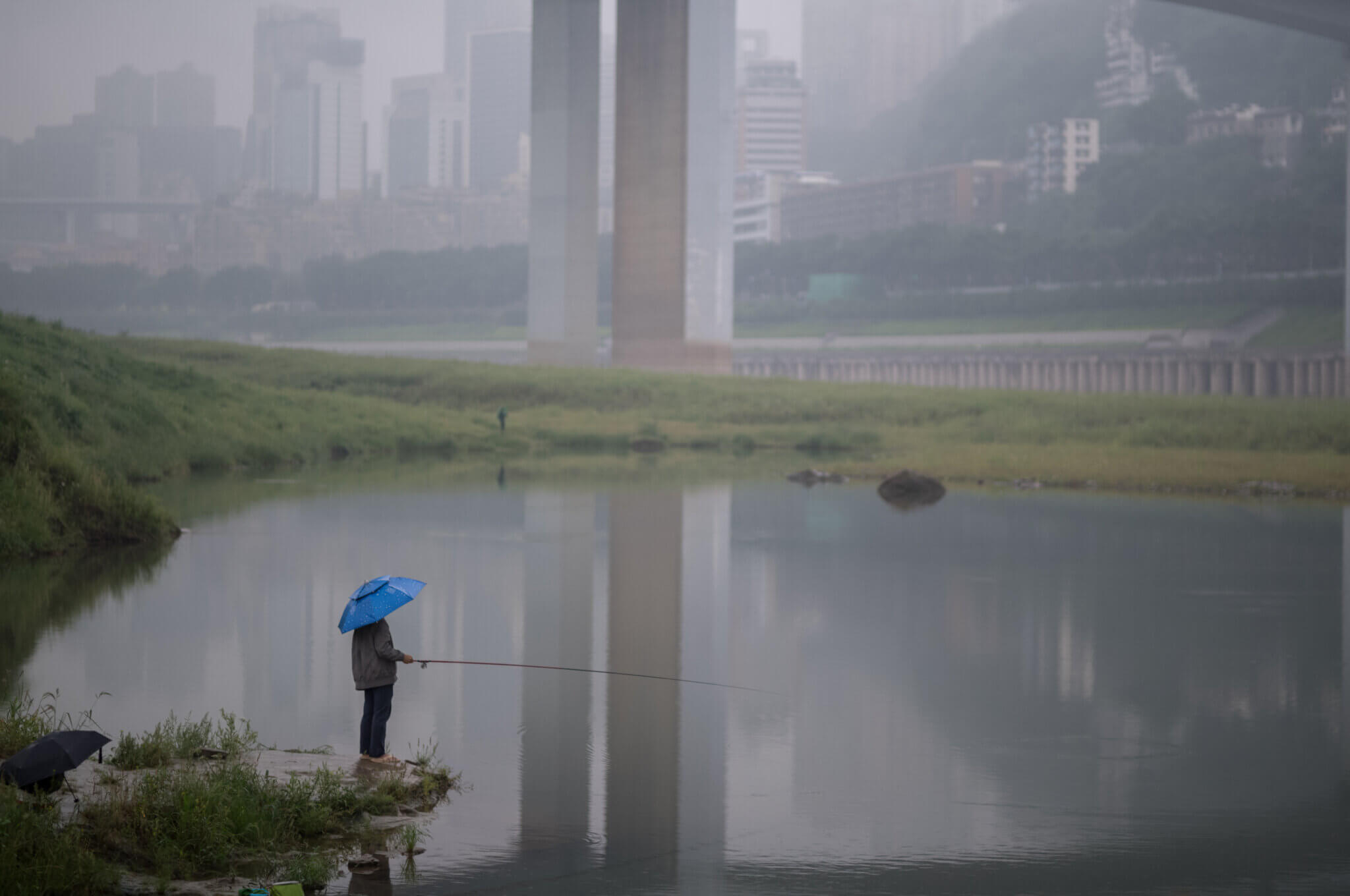 Lone man fishing on the shore of a body of water, with a sprawling city in the background.